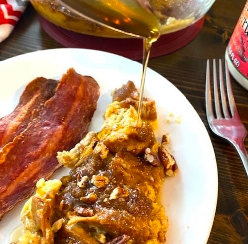 drizzling syrup over baked french toast casserole beside a couple of strips of bacon