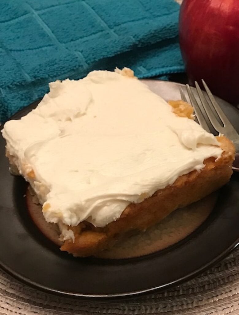 slice of frosted 2-ingredient apple cake on a plat beside an apple and a dishtowel